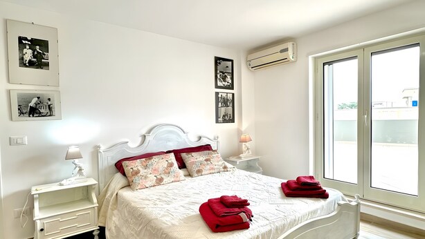 A white bedroom with a bed, a nightstand, a lamp, a rug, and wall-mounted pictures. - Apartment 'La Mansarda - Terrazze Relax A Due' with Balcony, Wi-Fi and Air Conditioning (Cellamare)