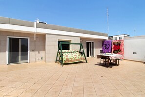 Un espace patio avec une balançoire, une table recouverte dâune nappe et un bĂątiment dotĂ© dâun sol carrelĂ© et dâune porte.