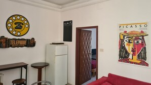 Une pièce avec un réfrigérateur blanc, une porte en bois, un canapé rouge, une petite table ronde et un téléphone fixé au mur.