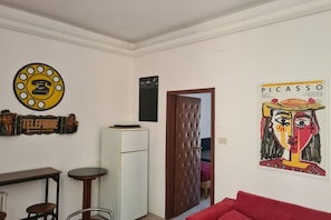Ein Zimmer mit einem weißen Kühlschrank, einer Holztür, einem roten Sofa, einem kleinen runden Tisch und einem an der Wand befestigten Telefon.