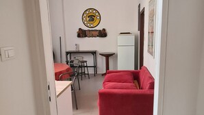 Un espace de vie compact comprenant un canapé rouge, une petite table à manger avec des chaises, un réfrigérateur blanc et un téléphone fixé au mur.