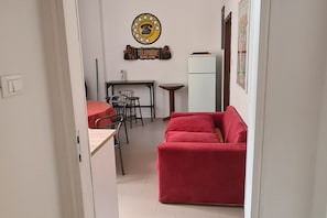 Ein kompakter Wohnraum mit einem roten Sofa, einem kleinen Esstisch mit Stühlen, einem weißen Kühlschrank und einem an der Wand befestigten Telefon.