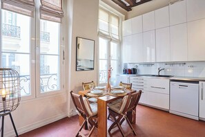 Ein Essbereich mit Tisch für vier Personen, eine Küche mit weißen Schränken und große Fenster mit Jalousien.
