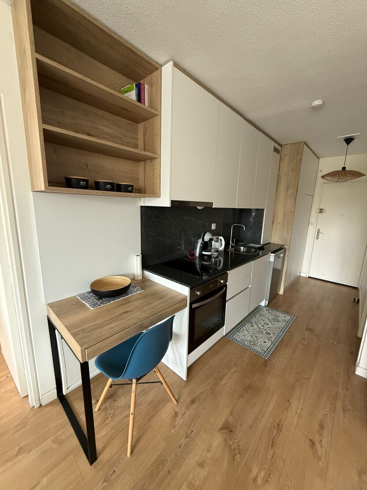 A compact kitchen with a wooden table, a blue chair, and a built-in shelf with books and containers.