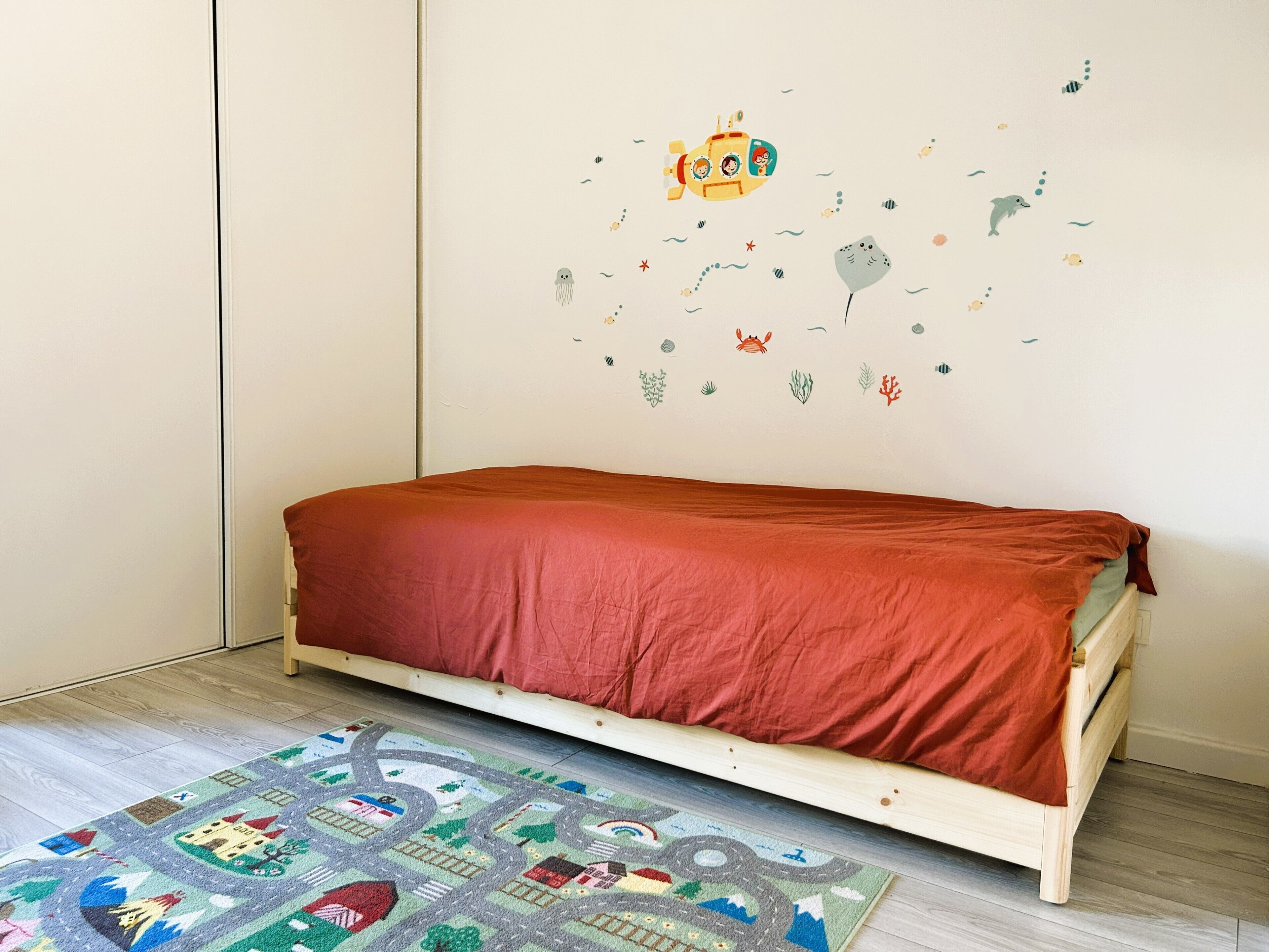 A single bed with a red blanket in a room with a white wall and a colorful rug.
