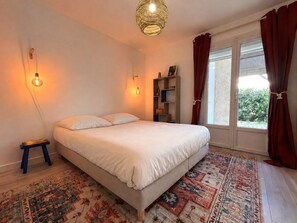 Ein Schlafzimmer mit einem Bett, zwei Wandleuchten, einem kleinen Nachttisch, einem Bücherregal und einem großen Fenster mit Vorhängen.