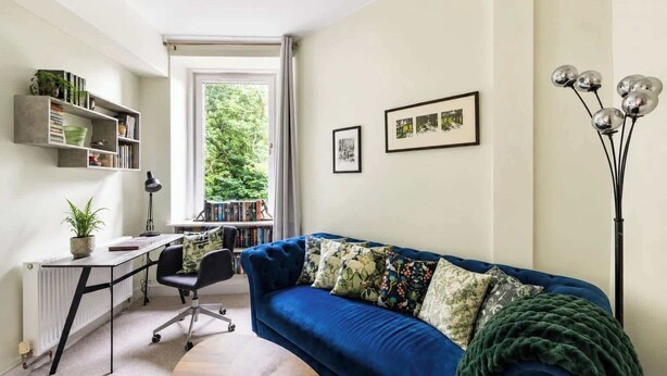 Una sala de estar moderna con un sofá azul, un escritorio con una silla, un radiador y un ventanal con vistas a la vegetación.