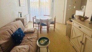 Un salon comprenant un canapé, une petite table ronde, un ventilateur de plafond et un coin cuisine avec des placards.