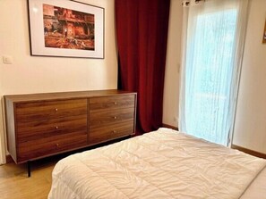 A bedroom with a bed, a wooden dresser, a framed picture on the wall, and a window with curtains. - Seaside apartment with terrace and parking (Aytré)