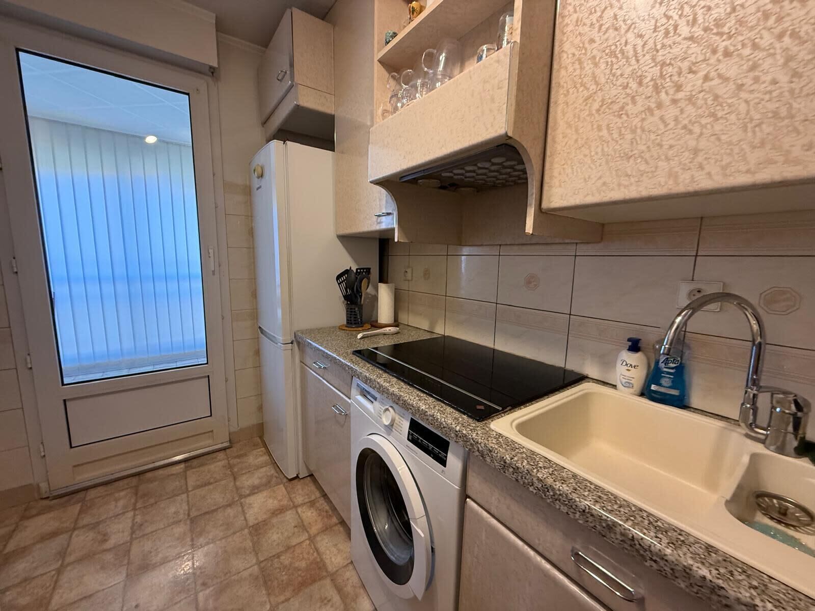 A compact kitchen with a washing machine, a built-in oven, a sink, and a window with blinds.