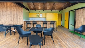 Un área de comedor al aire libre con techo, mesas y sillas, una cocina al fondo y un muro de bambú.