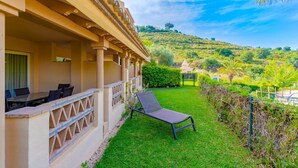 A hotel with a covered outdoor seating area, a lawn, and a view of a hill with greenery and a small structure. - GuestReady - Santa Maria Village (Marbella)