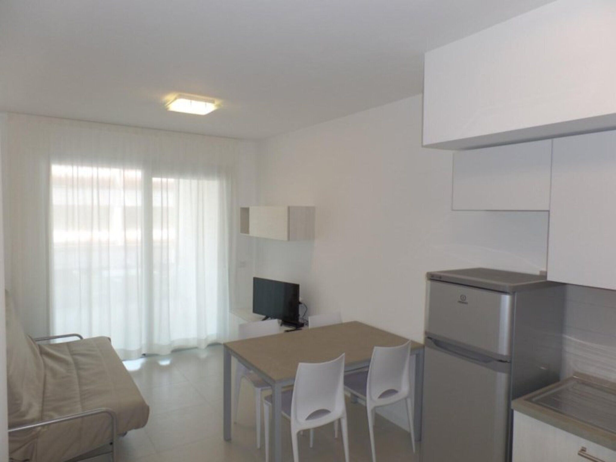 A compact kitchen with a refrigerator, a dining table with chairs, a TV, and a sofa in a bright room with a large window.