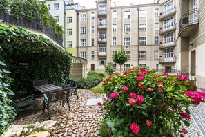 Ein Innenhof mit einer Steinterrasse, einer Bank und einer Hecke mit rosa Blüten.
