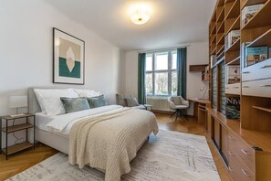 Ein Schlafzimmer mit Bett, Nachttischen, einem Sessel, einem Heizkörper, einem Fenster mit Vorhängen und einem Bücherregal.
