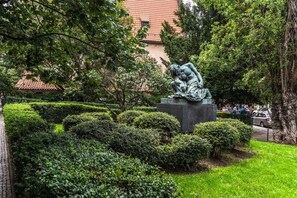 Eine Statue in einem gepflegten Garten mit geschnittenen Hecken und ein Gebäude mit rotem Ziegeldach im Hintergrund.