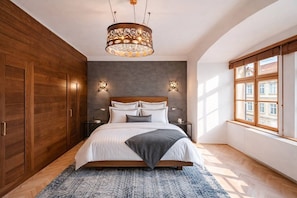 Ein Schlafzimmer mit einem großen Bett, einem Holzschrank, zwei Nachttischen, einer Kristallleuchtre und einem Fenster mit Blick auf Gebäude.