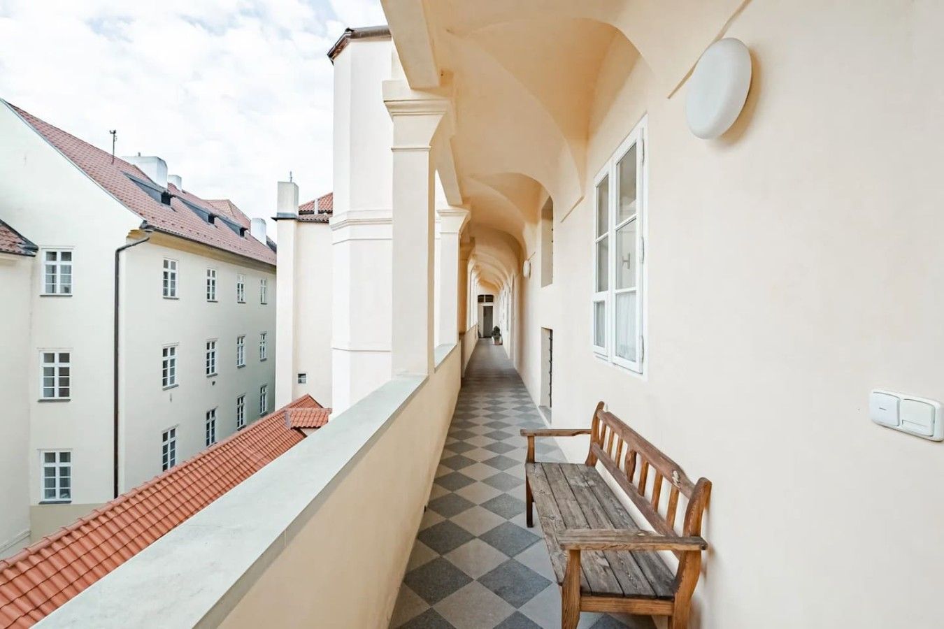 A corridor with a checkered floor, a wooden bench, and a view of adjacent buildings.