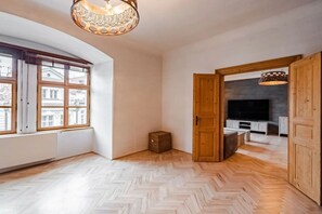 A spacious room with a herringbone floor, a large window with multiple panes, and a wooden door leading to another room with a television. - Luxury Historic Apartment | Next to Charles Bridge (Prague 1)