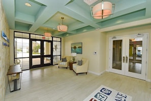 A spacious lobby with a high ceiling, a large glass door leading outside, and a seating area with two armchairs and a small table.