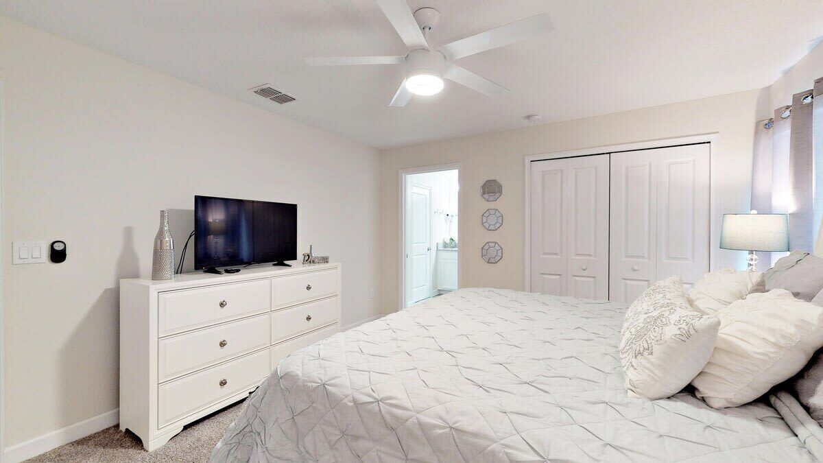 A bedroom with a bed, a TV on a dresser, a ceiling fan, and a door leading to another room.