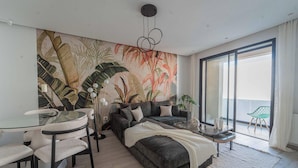 A modern living room with a large tropical-themed mural, a glass dining table with chairs, a sofa with cushions, and a small side table. - New 2BR Apt Parking Netflix La Marsa (La Marsa)