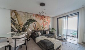 Ein modernes Wohnzimmer mit einer großflächigen Wandmalerei im tropischen Stil, einem Glastisch mit Stühlen, einer Couch mit Kissen und einem kleinen Beistelltisch.
