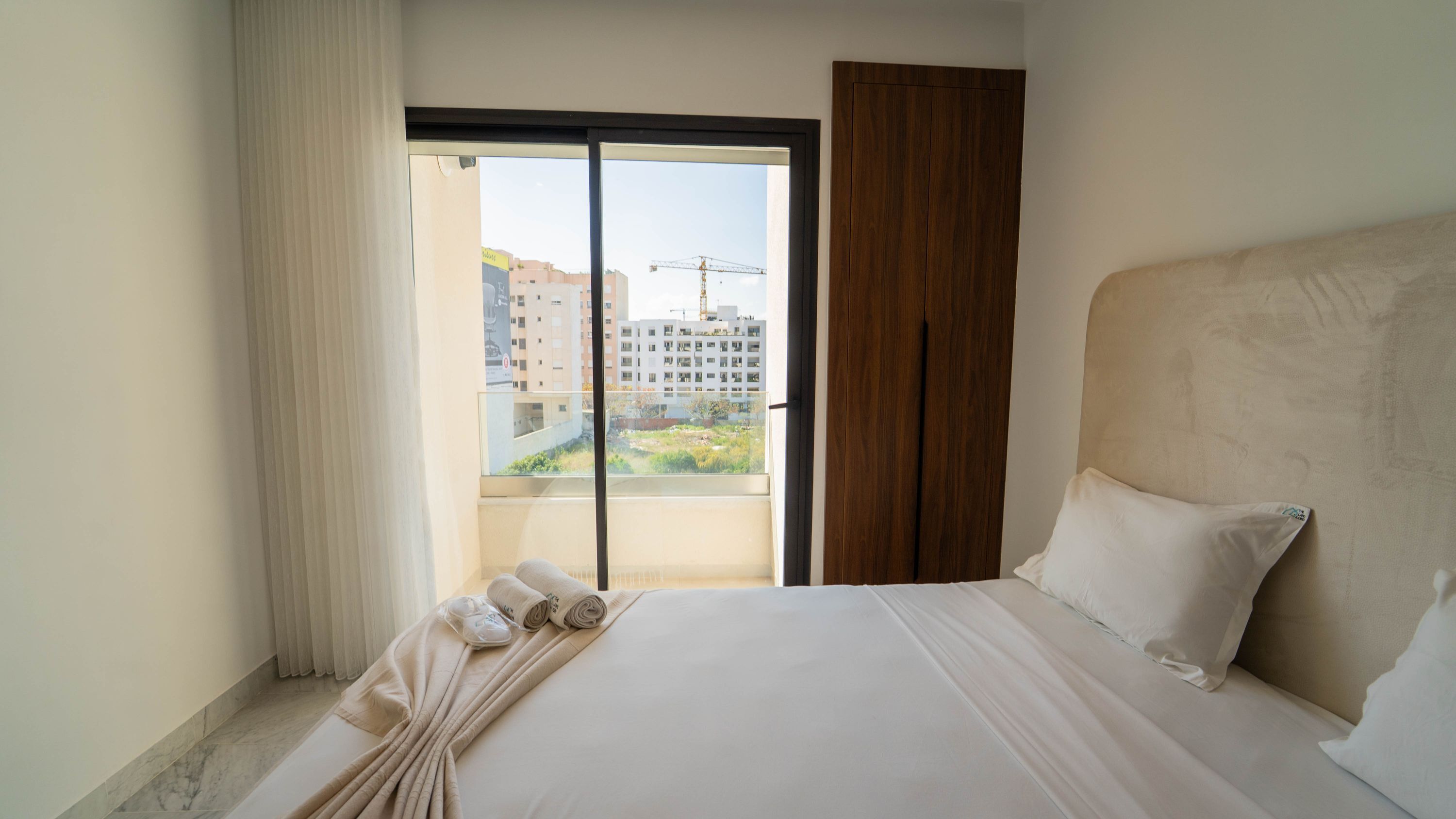 A hotel room with a large bed, a wooden headboard, a sliding glass door to a balcony, and a view of buildings and a crane.