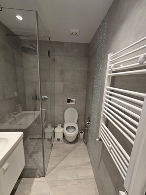 A modern bathroom with a glass shower enclosure, a white toilet, and a built-in towel rack. - Chic Boho 1BR Modern Comfort (La Marsa)