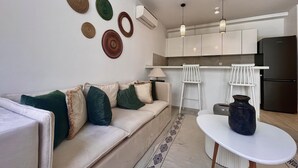 A modern living room with a sofa, a coffee table, and a kitchen area with cabinets and a refrigerator. - Chic Boho 1BR Modern Comfort (La Marsa)