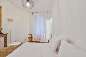 Ein Schlafzimmer mit Bett, Stuhl, Nachttisch, Lampe und einem Fenster mit Vorhängen.