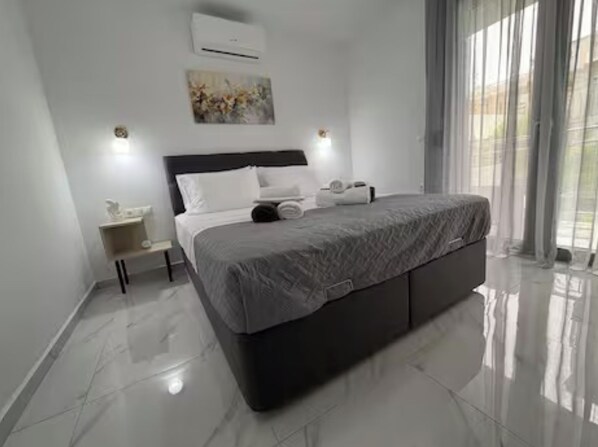 A modern bedroom with a large bed, a nightstand, an air conditioning unit, and a window with curtains.