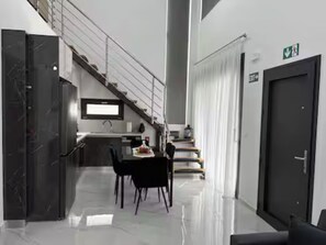 A modern interior with a dining area, kitchen, staircase, and a door with an emergency exit sign.