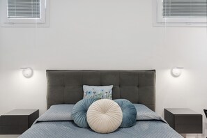 Een moderne slaapkamer met een grijs gestoffeerd hoofdbord, een bed met blauwe en beige beddengoed, twee nachtkastjes en wandlampen.