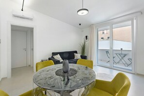 Een moderne woonkamer met een glazen tafel, gele stoelen, een zwarte bank en een schuifpui naar een buitenruimte.