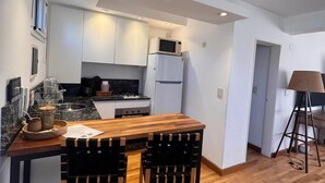 Une cuisine compacte avec des meubles blancs, un four à micro-ondes, un réfrigérateur, une table à manger en bois et deux chaises.