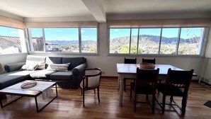 Un salon spacieux comprenant un canapé, une table à manger avec des chaises, un fauteuil en osier et de grandes fenêtres offrant une vue sur la montagne.