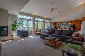 A spacious living room with a fireplace, large windows, a ceiling fan, and a modern kitchen in the background.
