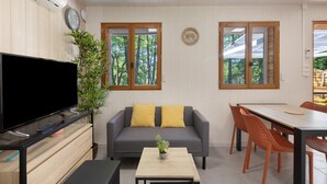 Een moderne woonkamer met een flatscreen televisie, een grijze bank met gele kussens, een eettafel met stoelen en grote ramen met uitzicht op bomen.
