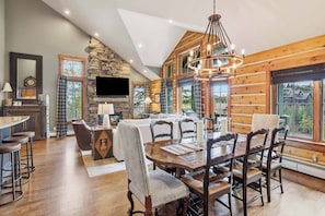 A spacious living area with a high ceiling, wooden walls, a stone fireplace, a dining table with chairs, and a large window offering a view of the outdoors. - Buckhorn Townhome 264 - 5 Bedroom (Beaver Creek)