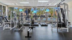 Un gimnasio con diferentes máquinas de ejercicio, ventanas amplias y vistas a palmeras y una piscina.