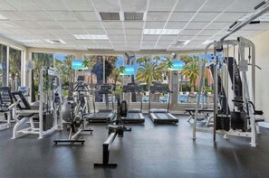 Ein Fitnessstudio mit verschiedenen Trainingsgeräten, großen Fenstern und Blick auf Palmen und einen Pool.