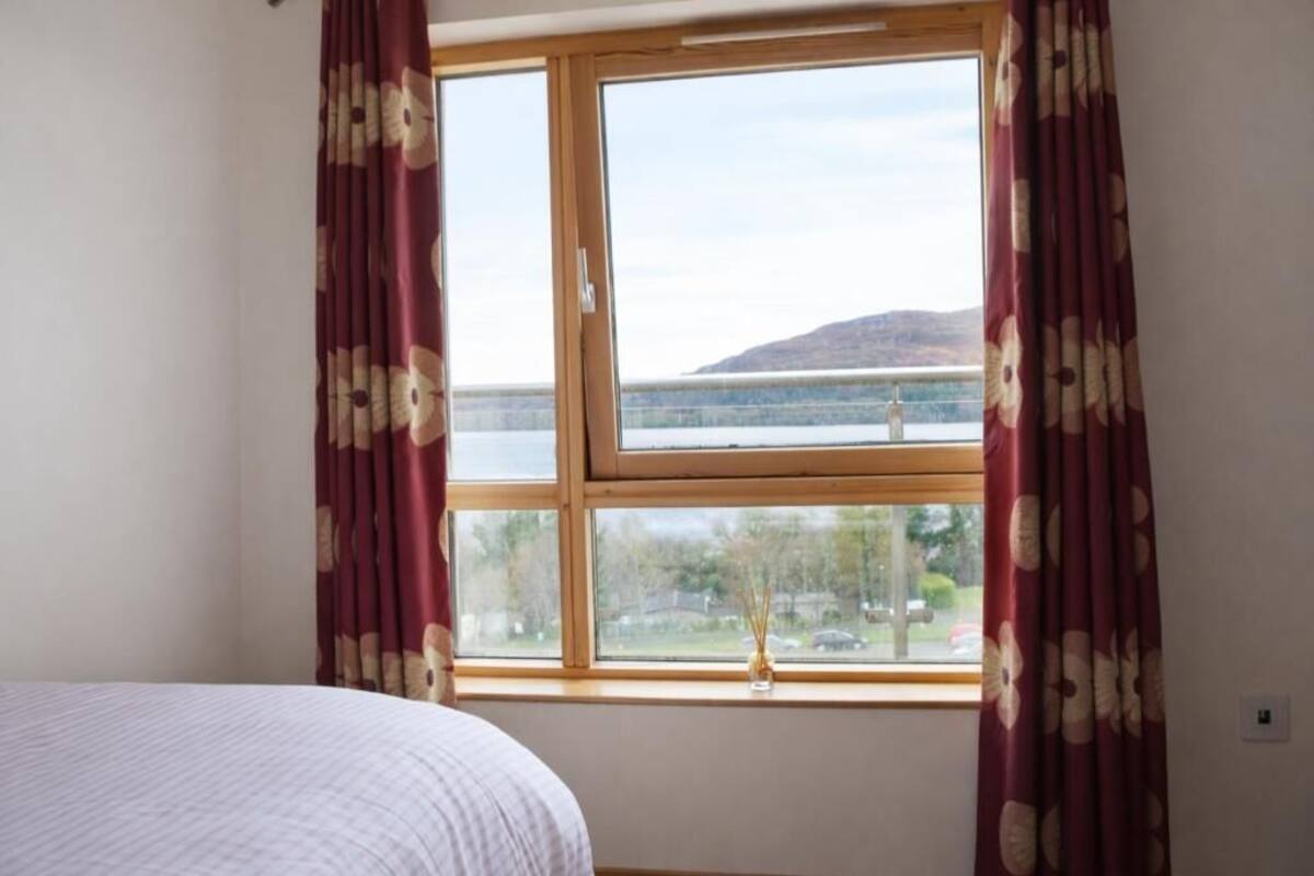 Ein Zimmer mit Bett, einem Vorhangfenster und Blick auf ein Gewässer und Hügel.