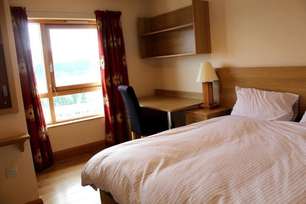 Ein Schlafzimmer mit Bett, Schreibtisch, Stuhl, Lampe und einem Fenster mit Vorhängen.