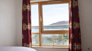 A room with a bed, a window with curtains, and a view of a body of water and hills.