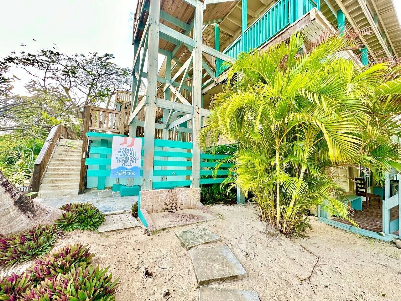 En villa vid stranden med en trappa som leder upp till en träveranda, omgiven av tropiska växter och en skylt som visar att den ligger vid stranden.