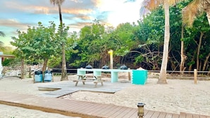 Zona arenosa de playa con una terraza de madera, mesas de picnic y una parrilla.