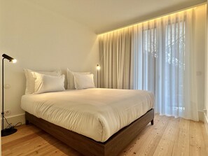 A bedroom with a wooden bed, white bedding, a bedside lamp, and sheer curtains. - Bolhão Prime | Luxe 3BR + Garage (Porto)