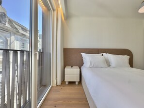 Habitación de hotel con un ventanal grande, cama con sábanas blancas, una mesita de noche y vistas a un edificio.