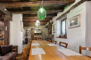 Un coin repas rustique avec une longue table en bois, des chaises, des lampes en verre vert suspendues, des murs en pierre et des poutres en bois.